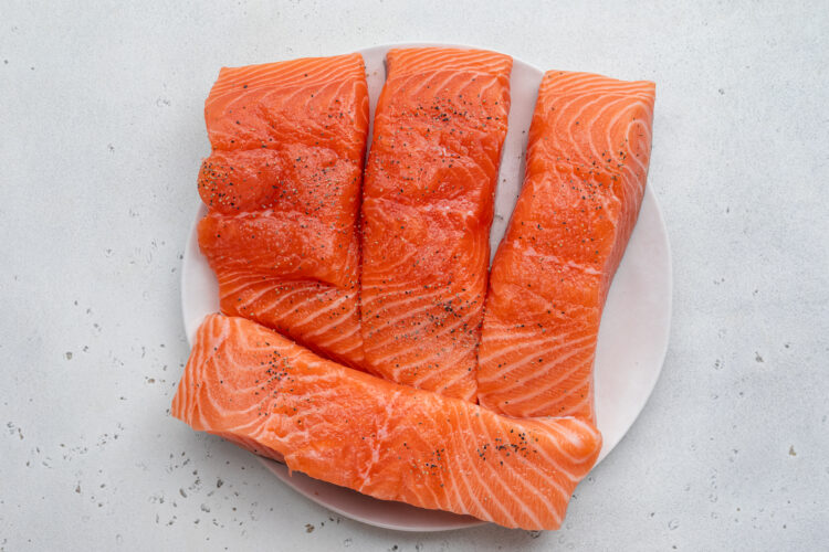 Overhead view of 4 uncooked, unseasoned, bright pink salmon fillets on a white plate on a white counter.