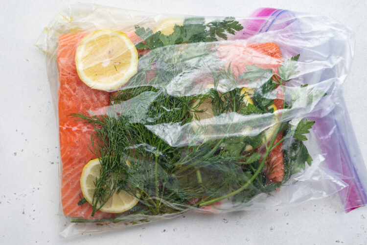 Overhead view of seasoned salmon fillets, lemon coins, and bright green fresh herbs sealed together in a Ziploc bag.