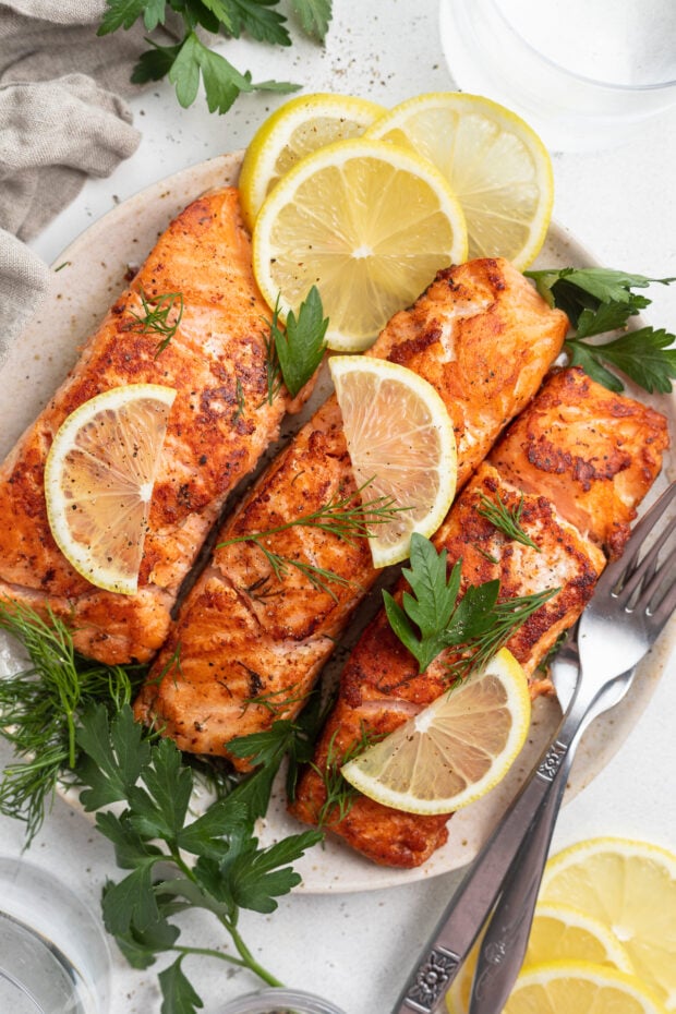 Overhead view of seasoned, pink, sous vide salmon on a white plate with lemon wedges and green garnishes.