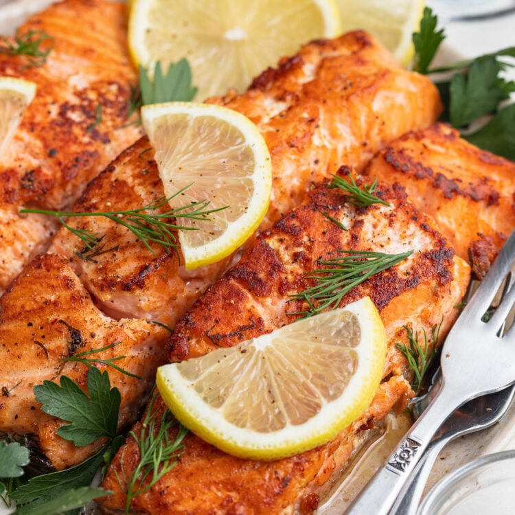 Angled, slightly zoomed out view of 3 seasoned sous vide salmon fillets on a white plate topped with lemon wedges and green parsley.