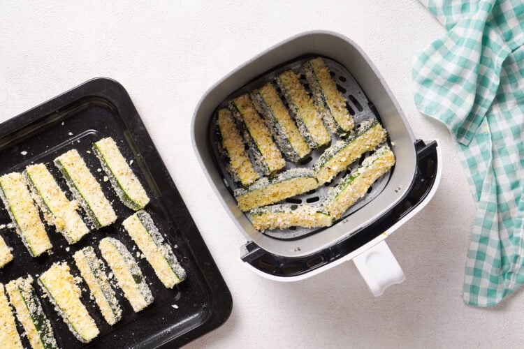 Overhead view of zucchini fries in air fryer basket next to a tray of uncooked zucchini fries.