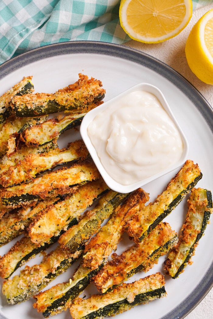 Overhead view of air fryer zucchini fries on a round plate with a square ramekin of garlic aioli.