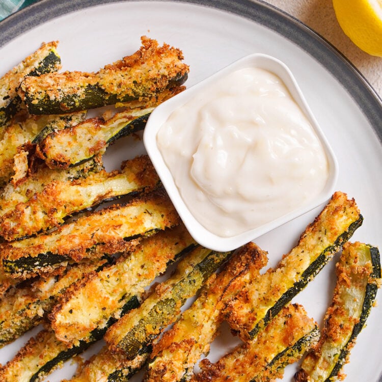 Overhead view of air fryer zucchini fries on a round plate with a square ramekin of garlic aioli.