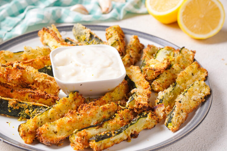 Side view of a platter of air fryer zucchini fries with a ramekin of garlic aioli on a table.