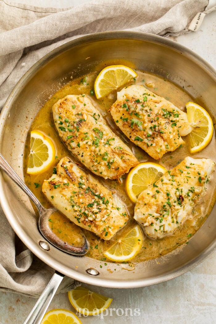 Overhead view of a skillet of cod fillets in a garlic butter sauce with lemon coins on a table.