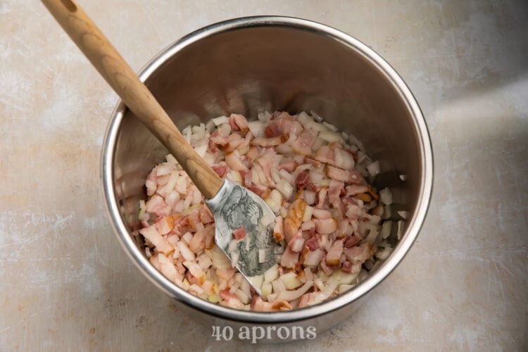 Overhead view of sauteed bacon and onions in an Instant Pot insert with a silicone spatula with a wooden handle.