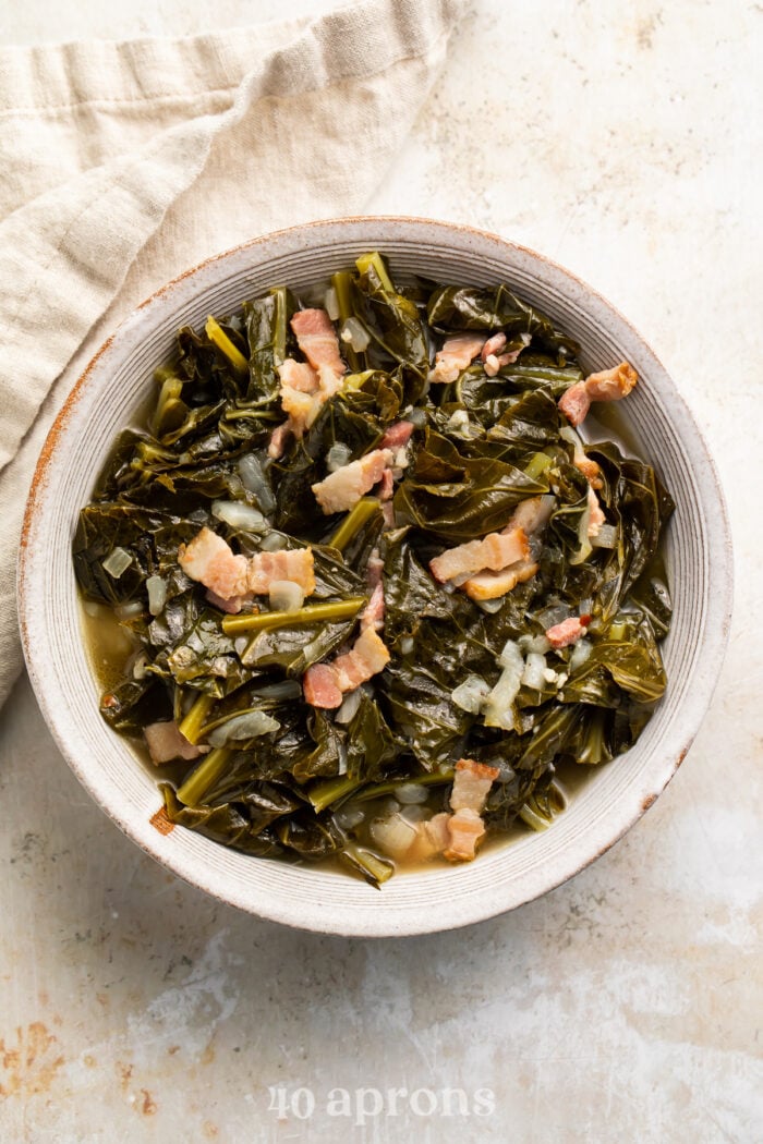 Overhead, zoomed out view of a vintage white bowl of collard greens with bacon on a neutral tabletop.
