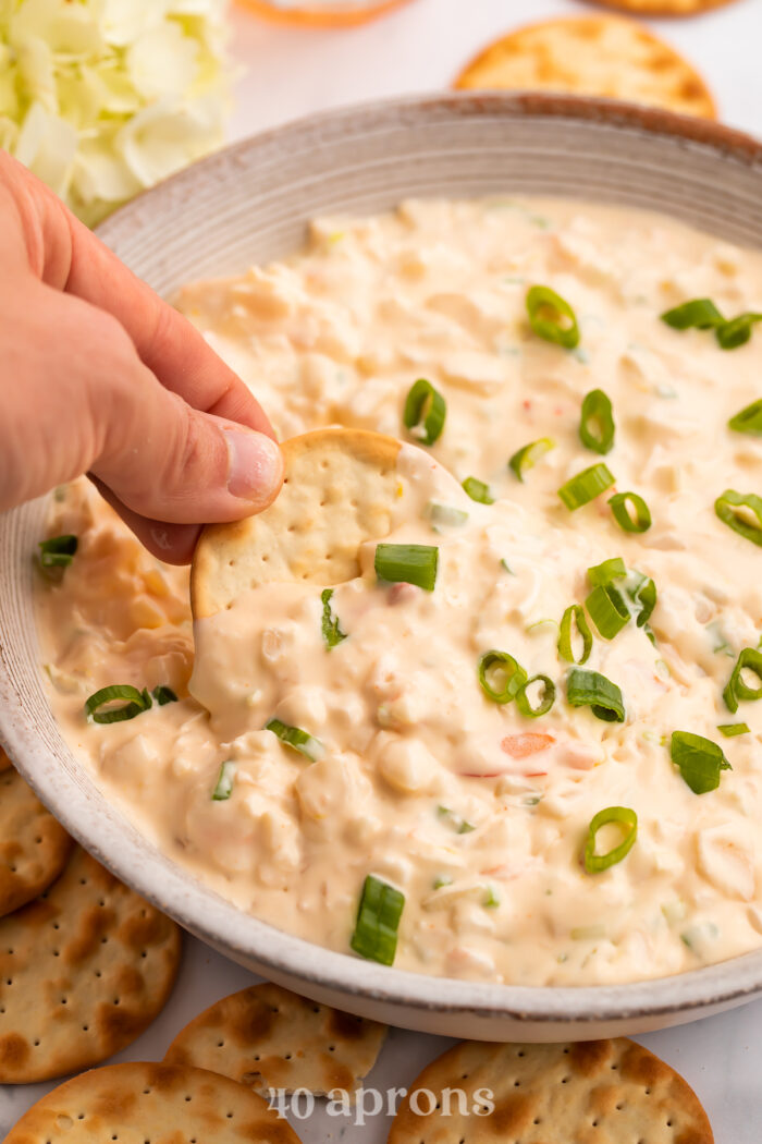 A hand holding a cracker, using it to scoop a helping of shrimp dip out of a large bowl.