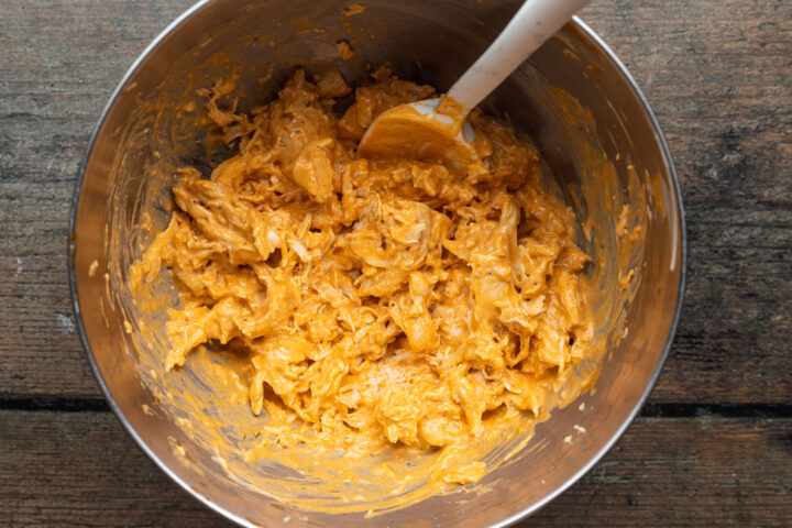 Overhead view of a large silver mixing bowl filled with shredded buffalo ranch chicken, with a white mixing spoon in the upper right corner.
