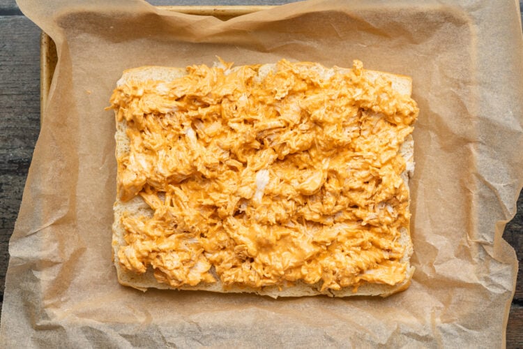 Overhead view of buffalo chicken spread out on top of a slab of halved sweet dinner rolls in a baking dish lined with parchment paper.