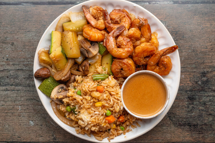 Overhead view of hibachi shrimp on a plate with fried rice, sautéed zucchini and mushrooms, and a small bowl of spicy mustard sauce.