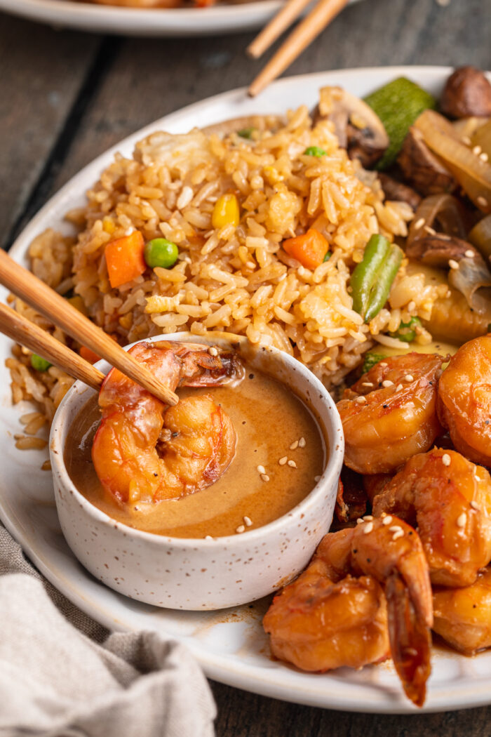 Image showing hibachi shrimp, held by chopsticks, being dipped into a small ramekin of mustard sauce on a plate with fried rice.