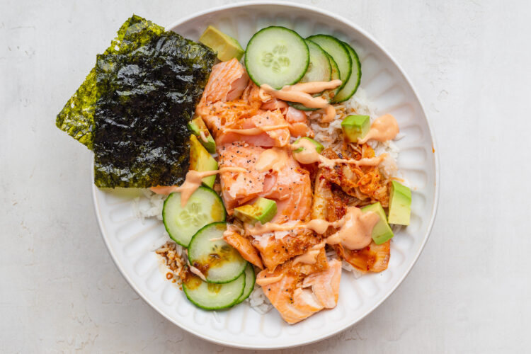 Overhead view of a salmon rice bowl with cucumber, avocado, kimchi, and nori, topped with sriracha mayo and soy sauce.