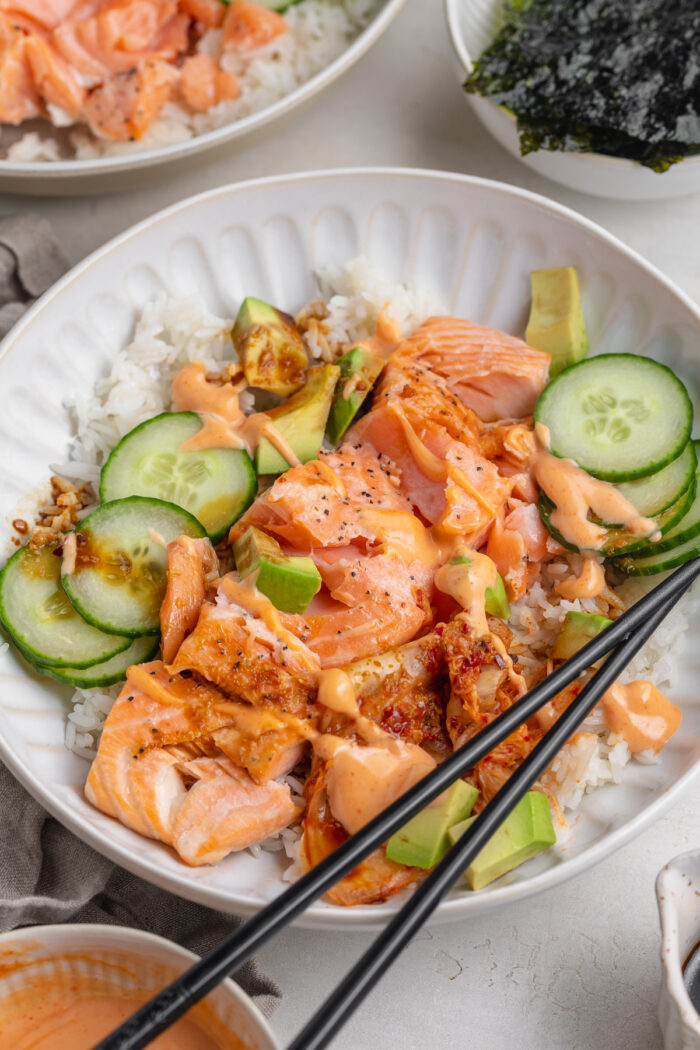 Angled view of a salmon rice bowl with salmon, rice, cucumber, avocado, seaweed, and chopsticks on a grey table.