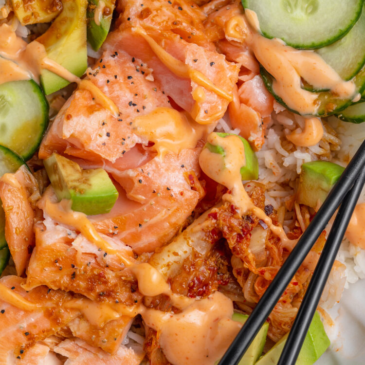 Close-up view of oven-baked salmon fillets with white rice, soy sauce, creamy sriracha mayo, cucumber, avocado, and chopsticks.