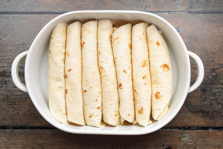 Overhead view of 7 rolled enchiladas in a white oval casserole dish on a neutral wooden background.