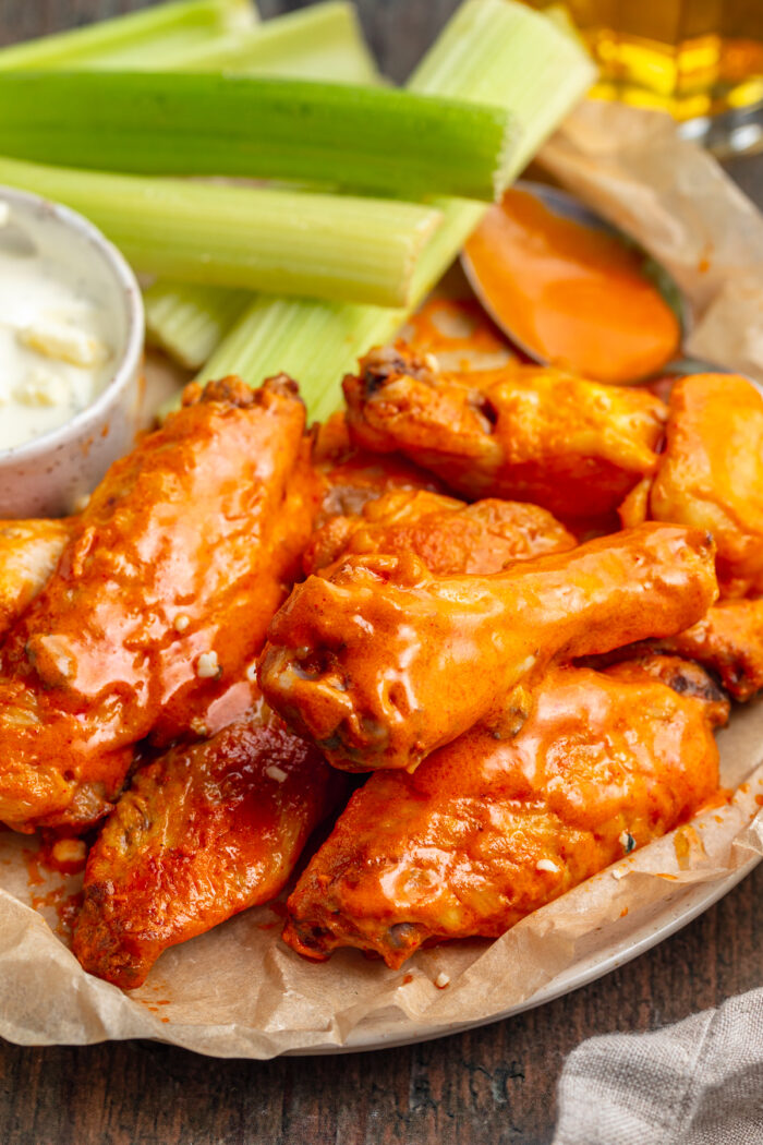 Overhead view of chicken wings coated in a glistening orange buffalo sauce on a sheet of tan parchment paper with bright green celery in the background.