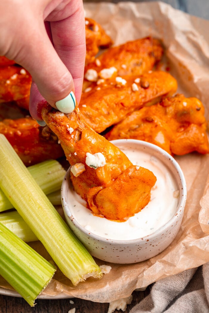 A white woman's hand dips an orange chicken wing in a small bowl of white ranch dressing that sits next to sticks of green celery.