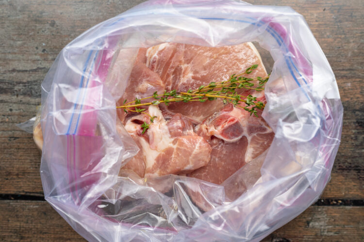 Overhead view of seasoned pork chops, sprigs of fresh herbs, and smashed garlic in a large Ziploc bag.