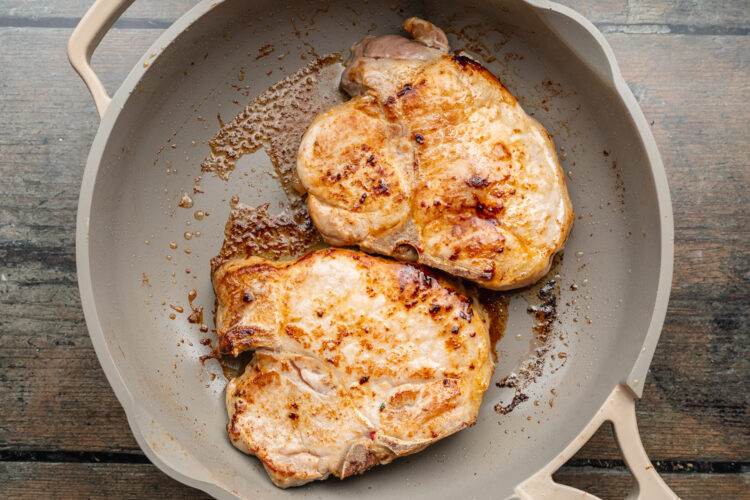 Overhead view of two pan-seared pork chops in a large skillet on a wooden tabletop.