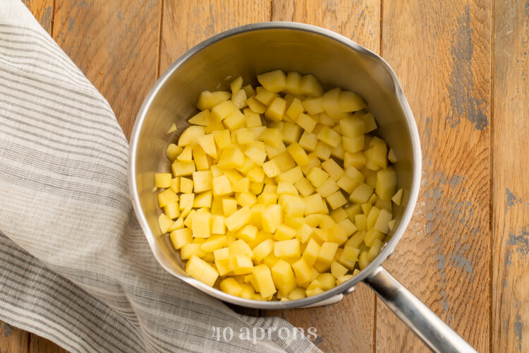 Overhead view of chopped sweet apples in a medium silver saucepan with handle, sitting on a wooden tabletop with a neutral napkin.