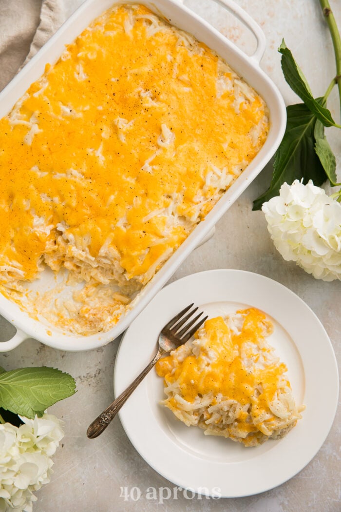 Overhead view of a cheesy copycat Cracker Barrel hashbrown casserole in a casserole dish, with a large scoop of casserole on a plate next to the dish.