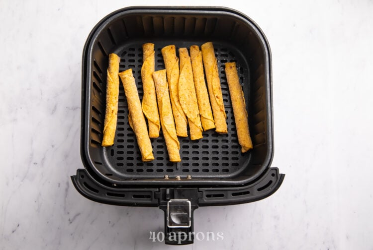 Overhead view of crispy frozen corn taquitos in an air fryer basket on a white countertop.