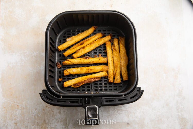 Overhead view of crispy cooked taquitos in an air fryer basket resting on a white and grey countertop.