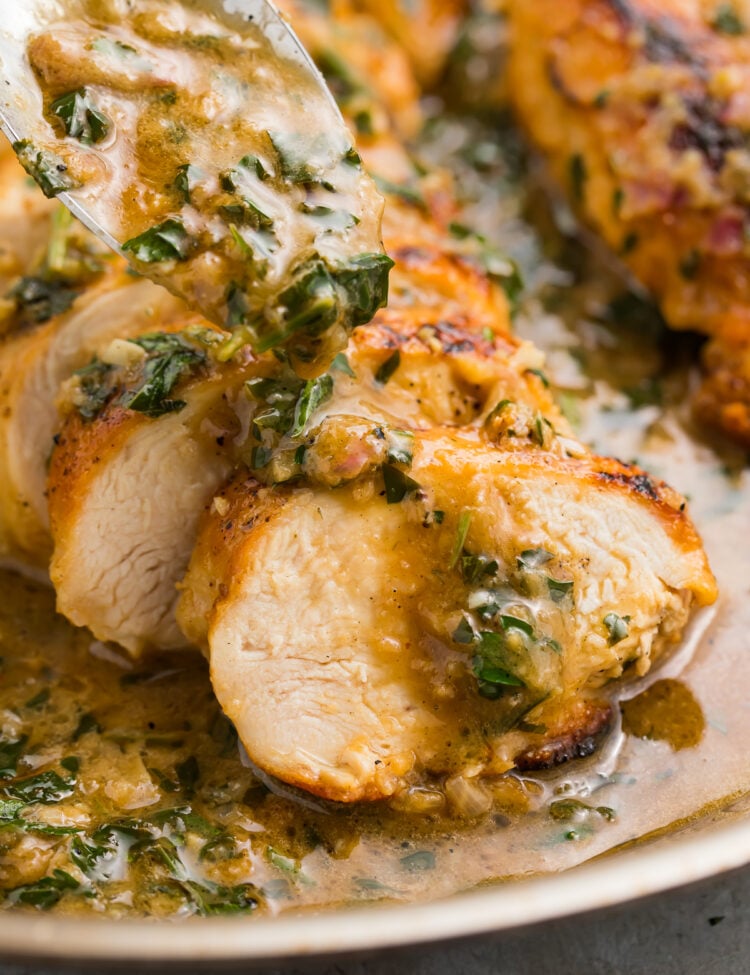 Close-up view of garlic herb butter sauce pouring from a spoon onto slices of tender chicken breast on a plate.
