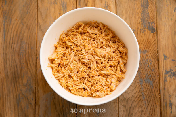 Overhead view of a white bowl containing orange shredded chicken on a light brown wooden table.