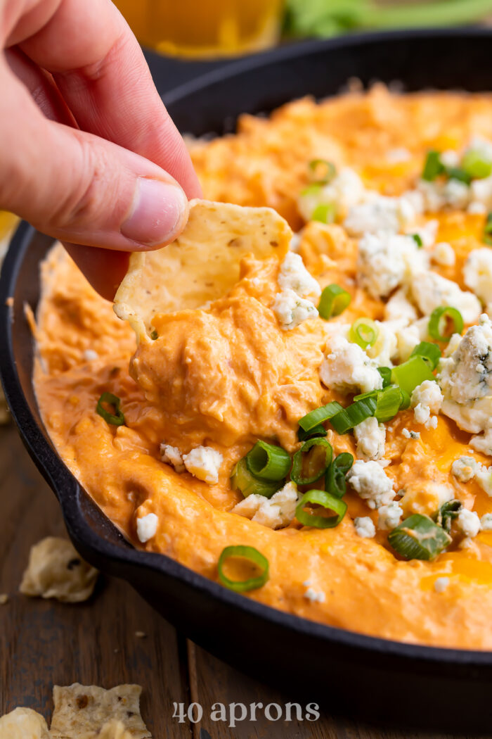 A white hand holding a tortilla chip scooping buffalo chicken dip topped with crumbled cheese and green onions out of a cast iron skillet.