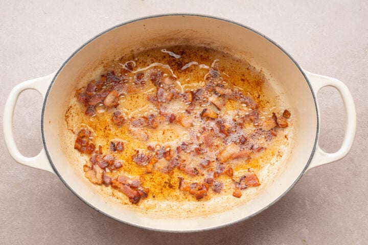 Overhead view of diced bacon strips cooked in bacon fat in the bottom of a large oval pot with handles.