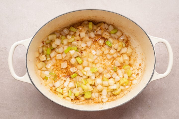 Overhead view of diced onion, chopped celery, and minced garlic in a large pot with rendered bacon fat on a neutral table.