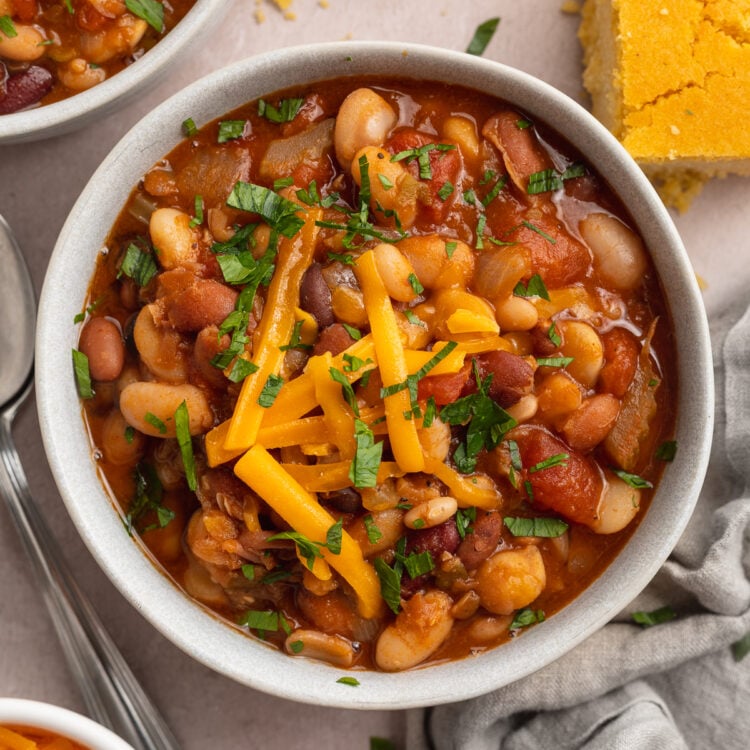 Overhead view of a bowl of deep red 15 bean soup topped with chopped green onions and yellow shredded cheddar in a white bowl on a white table with spoons, chopped onions, and cornbread.