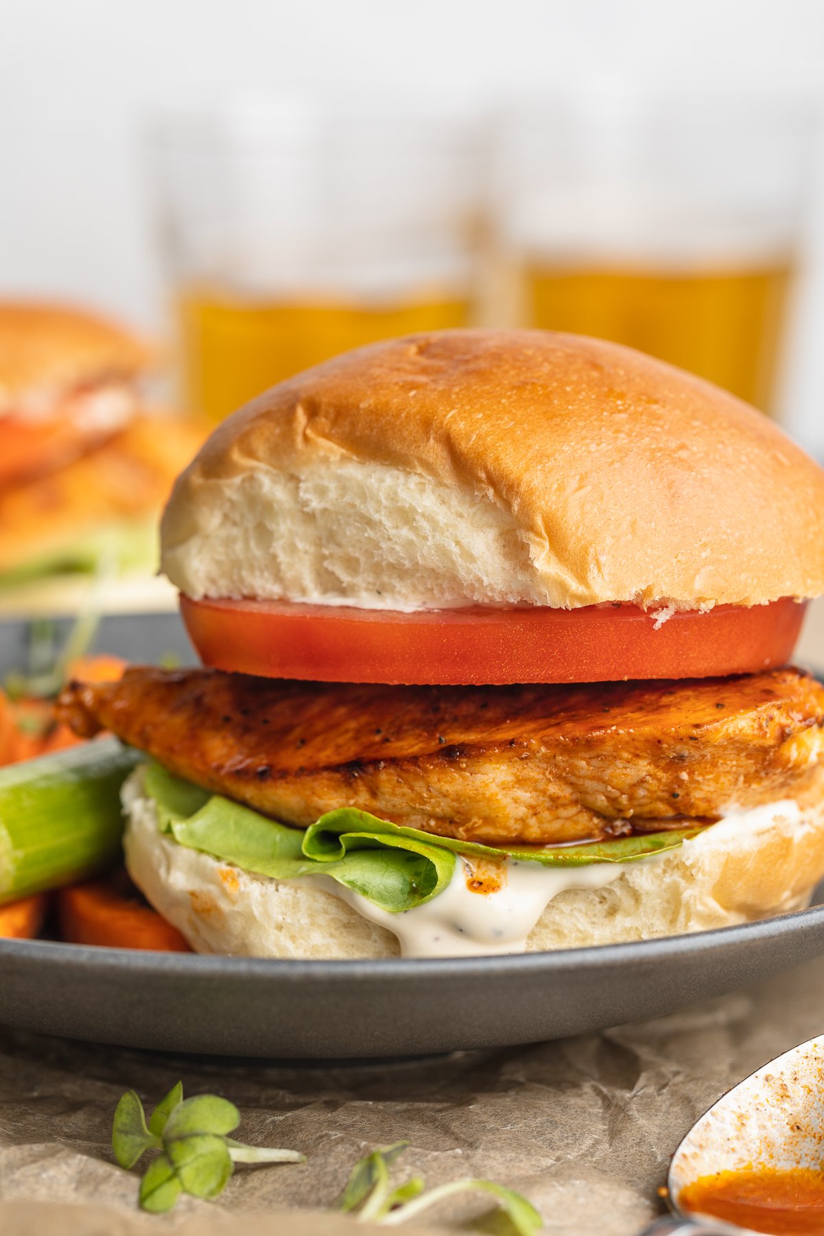 Side view of a buffalo chicken sandwich with an orange-colored grilled chicken breast, a thick slice of tomato, green lettuce, and a fluffy bun on a blue-grey plate.