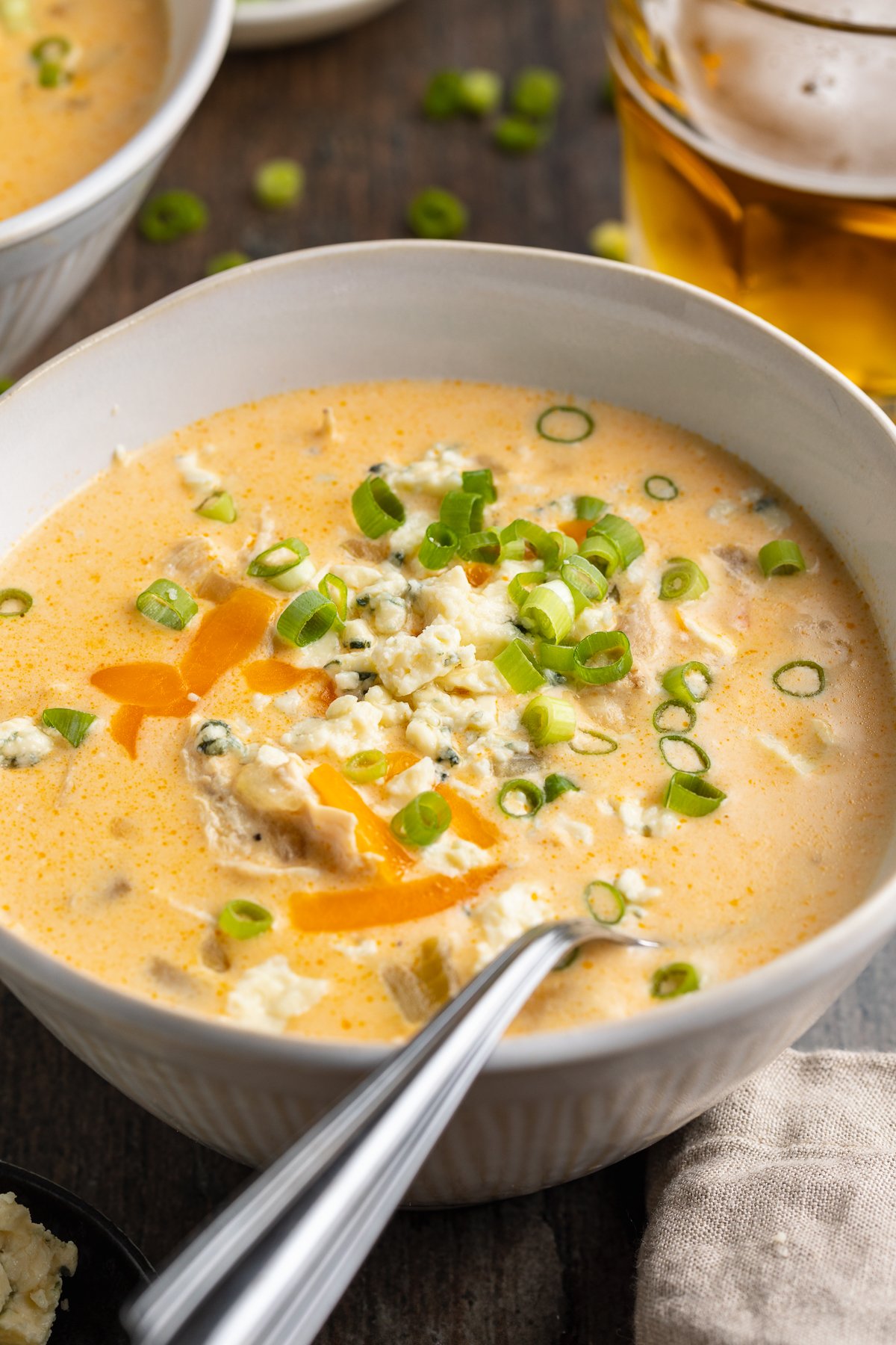 Angled view of a misshapen white bowl containing pale orange buffalo chicken soup topped with green onions and cheese.