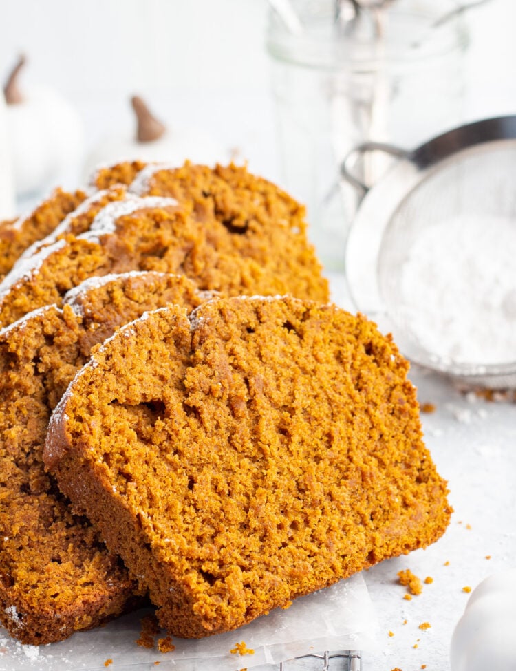 Side view of bright orange slices of gluten-free pumpkin bread topped with powdered sugar leaning up on one another on a white tabletop.