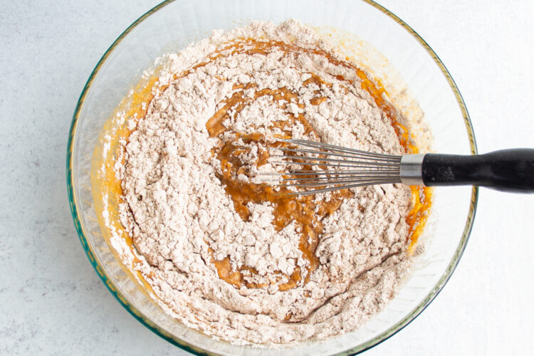 Overhead view of dry bread ingredients add to large glass mixing bowl with wet bread ingredients and incorporated with a whisk.