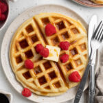 Overhead view of a plate of round, fluffy, Belgian-style gluten-free waffles topped with butter, syrup, and fresh red raspberries with silverware.