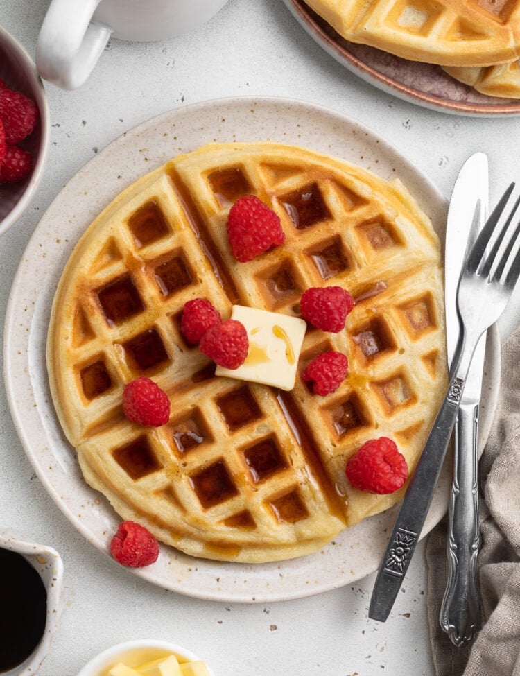 Overhead view of a plate of round, fluffy, Belgian-style gluten-free waffles topped with butter, syrup, and fresh red raspberries with silverware.