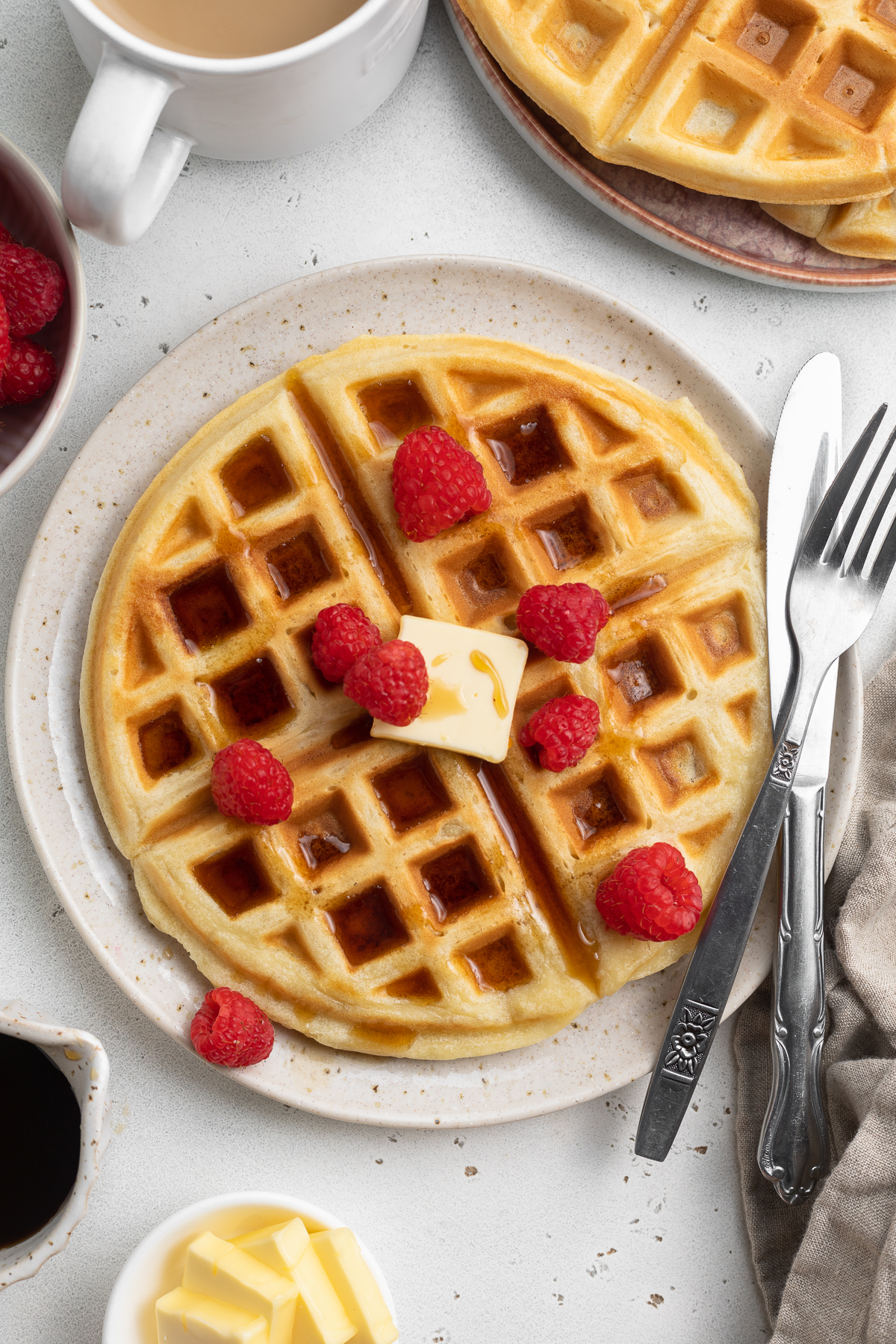 Overhead view of a plate of round, fluffy, Belgian-style gluten-free waffles  topped with butter, syrup, and fresh red raspberries with silverware.