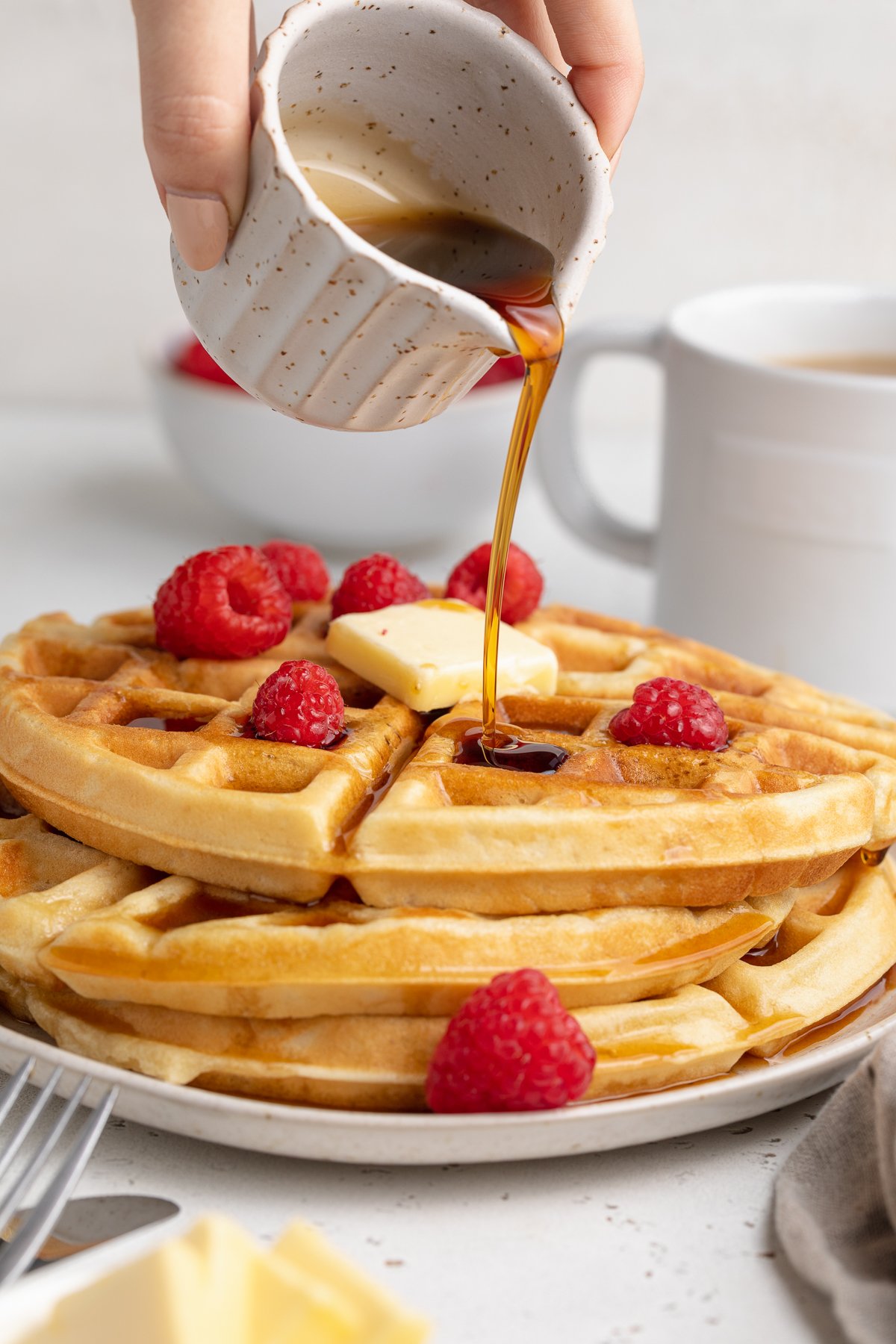 Side view of a stack of 3 round gluten-free waffles on a white plate. A hand pours syrup over the top of the waffles and onto butter and raspberries.