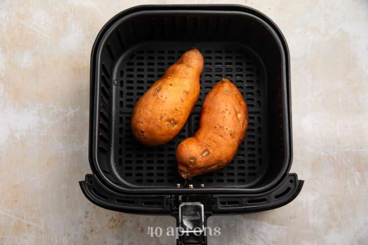 Overhead view of two medium sweet potatoes in a black air fryer basket on a neutral countertop.