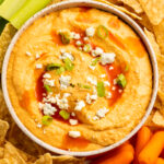 Close-up, overhead view of a bowl of buffalo hummus topped with blue cheese crumbles in the center of a platter surrounded by celery, carrots, and tortilla chips.