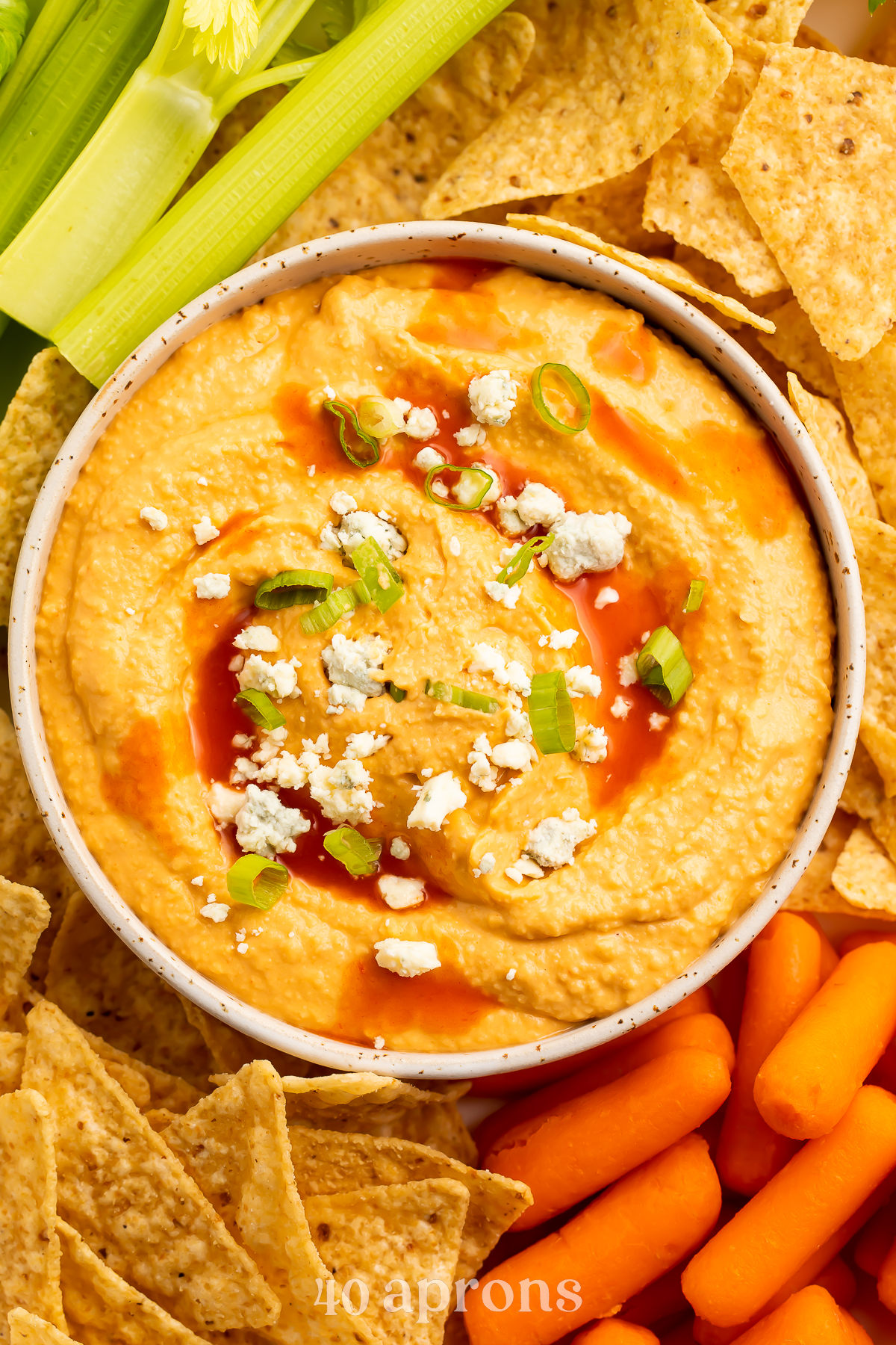 Close-up, overhead view of a bowl of buffalo hummus topped with blue cheese crumbles in the center of a platter surrounded by celery, carrots, and tortilla chips.