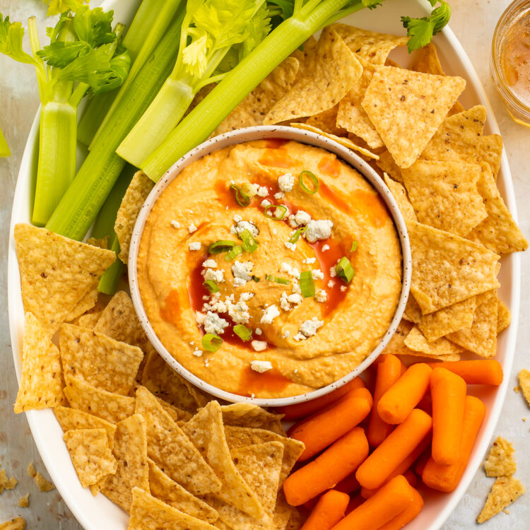 Overhead view of a bowl of buffalo hummus topped with blue cheese crumbles in the center of a platter surrounded by celery, carrots, and tortilla chips.