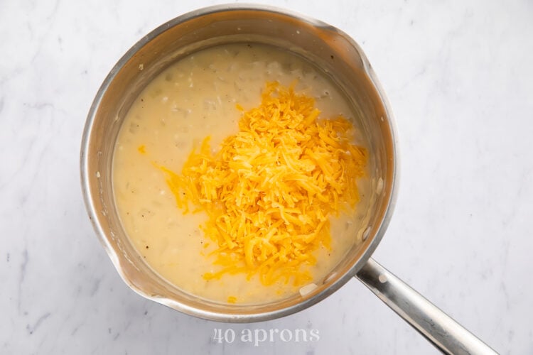 Overhead view of chicken broth, garlic, onions, flour, and shredded cheddar cheese in a medium silver saucepan with a silver handle.