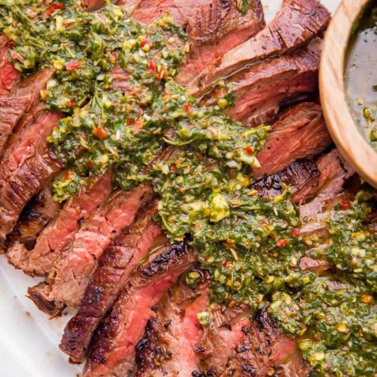 Close-up square photo of seared, medium-rare flank steak topped with bright green chimichurri sauce.