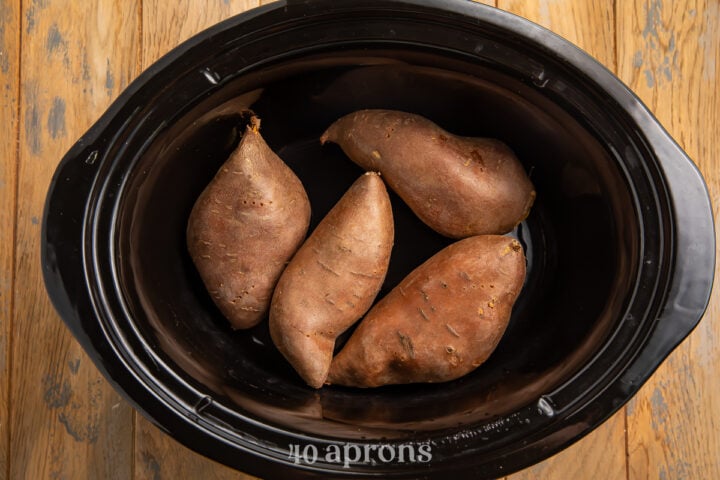 Overhead view of 4 large sweet potatoes in the bottom of a black slow cooker dish insert.