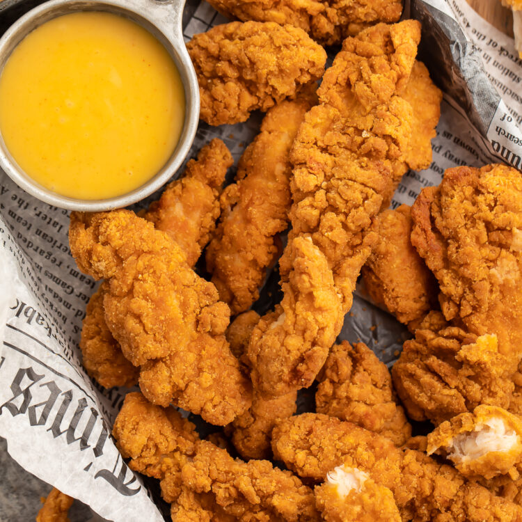 Close up overhead view of a pan lined with newspaper filled with chicken tenders and a small bowl of yellow honey mustard dipping sauce.
